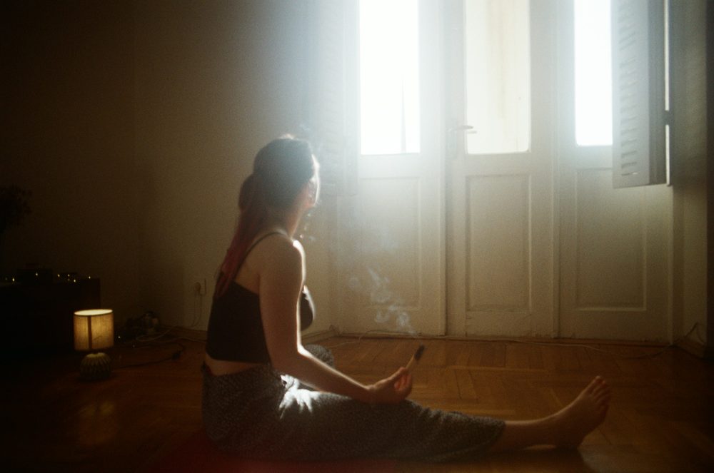 A person seated on the floor of a dim room, lit by daylight from a tall window.